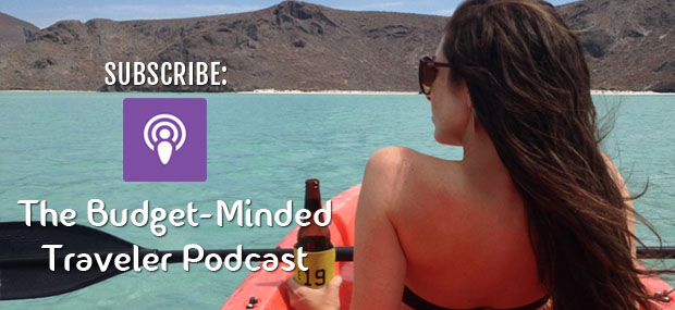 A woman with long brown hair and sunglasses sits in a red kayak, holding a beer bottle wrapped in a yellow koozie, while gazing at the turquoise water and rugged mountains in the background. Overlay text reads, "SUBSCRIBE: The Budget-Minded Traveler Podcast," with a purple podcast icon. The image evokes a sense of adventure and relaxation, promoting a travel-focused podcast.