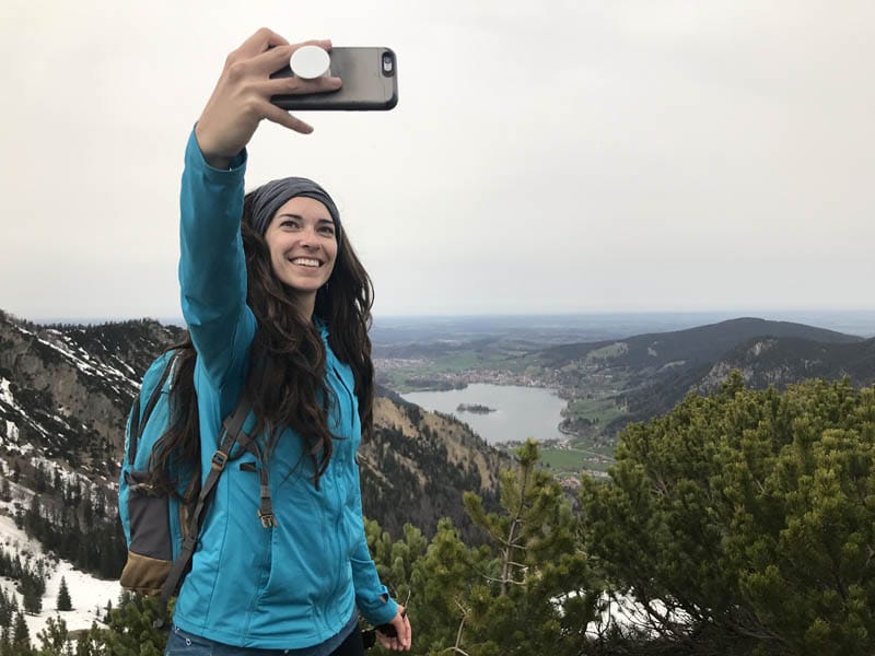 A smiling woman in a blue jacket takes a selfie on a mountain trail, with a scenic view of a valley and lake in the background, conveying a sense of adventure and the enjoyment of nature.