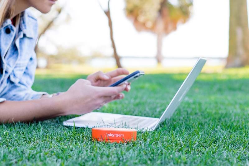 Outdoor work setup featuring a person sitting on the grass, using a smartphone with a laptop open nearby, and a Skyroam portable Wi-Fi hotspot device in the foreground, implying mobile connectivity in a park setting.