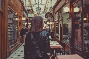 A woman in a black leather jacket walks through a charming, covered passageway lined with quaint shops and cafes. The scene exudes a vintage European vibe, with warm lighting and inviting storefronts.