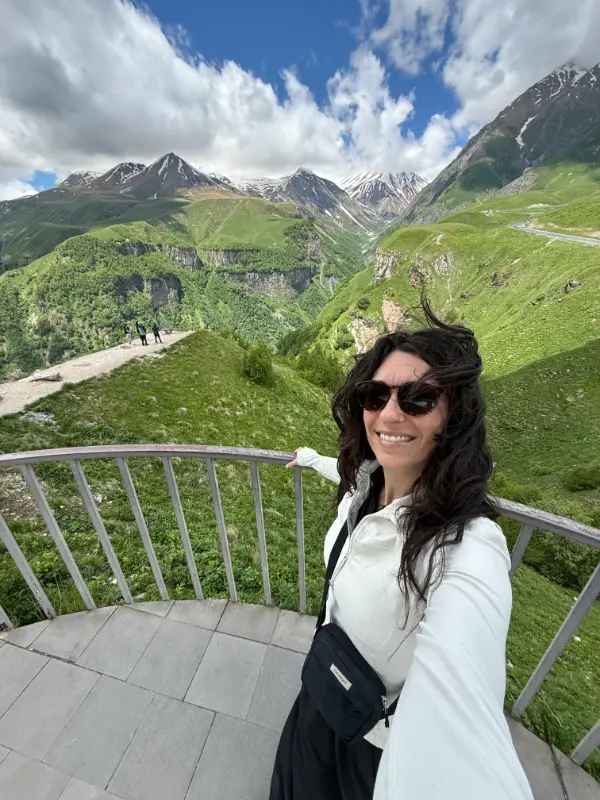 A woman in sunglasses and a white jacket takes a selfie from a mountain overlook with a black crossbody bag across her chest. Green valleys and steep snow streaked peaks stretch behind her beneath a bright blue sky with large white clouds.