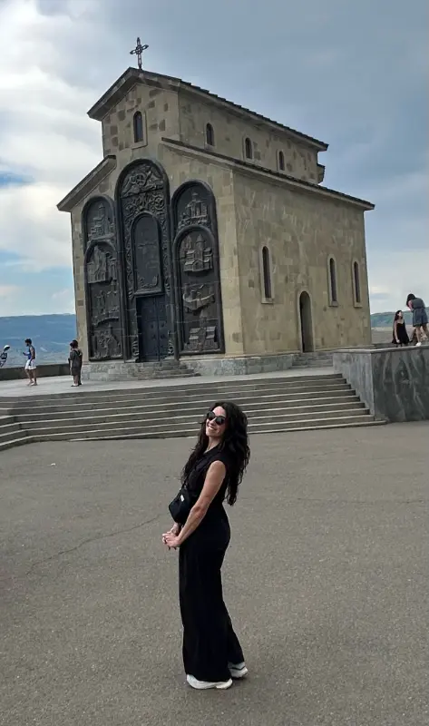A woman in black clothing and sunglasses poses in front of a stone church with tall ornate bronze doors and a cross on the roof. Wide steps lead up to the church while a few visitors stand around the plaza under a cloudy sky.
