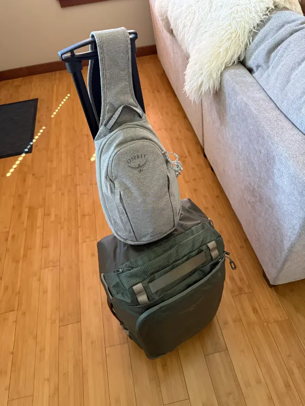 A gray Osprey sling bag is looped over the extended handle of a dark green rolling suitcase on a hardwood floor. The luggage sits beside a light gray sofa with a white textured pillow.