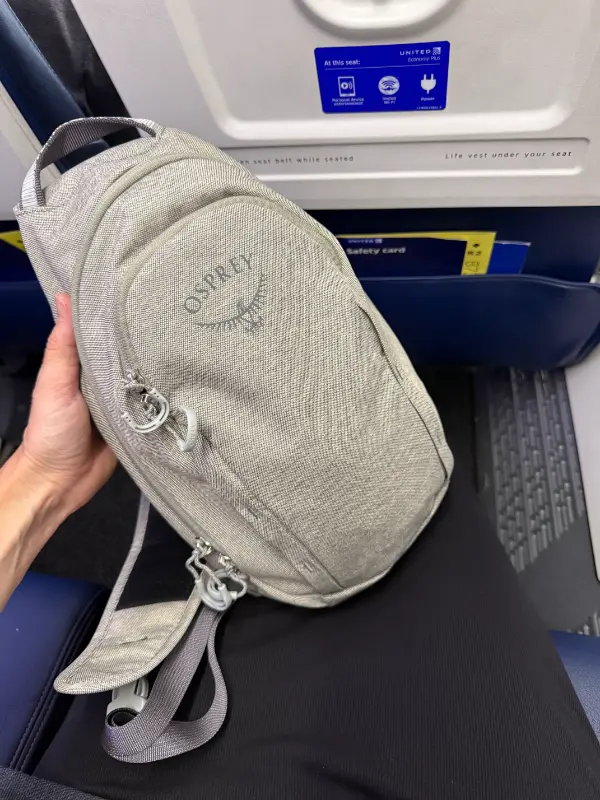 A gray Osprey sling bag rests on an airplane seat while being held up for the camera. The seatback in front shows United Economy Plus signage and a pocket holding the safety card.